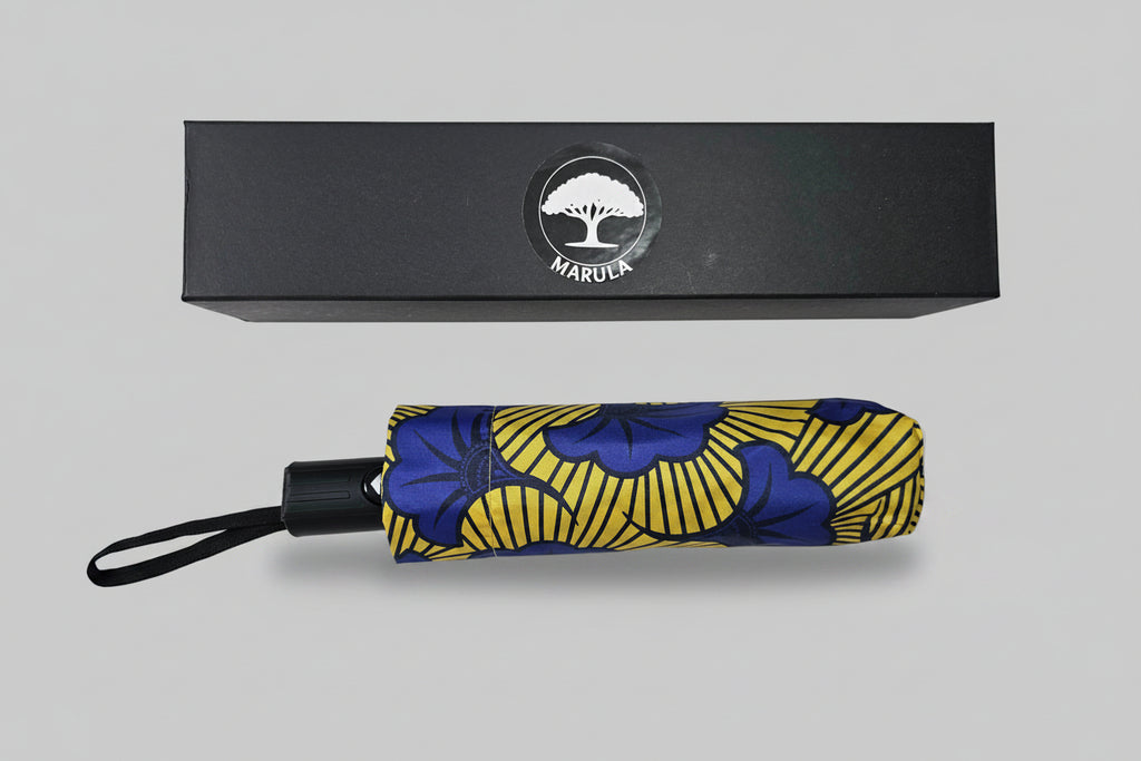Foldable umbrella with blue and yellow pattern and black handle, placed in front of a black box with 'Marula' branding.
